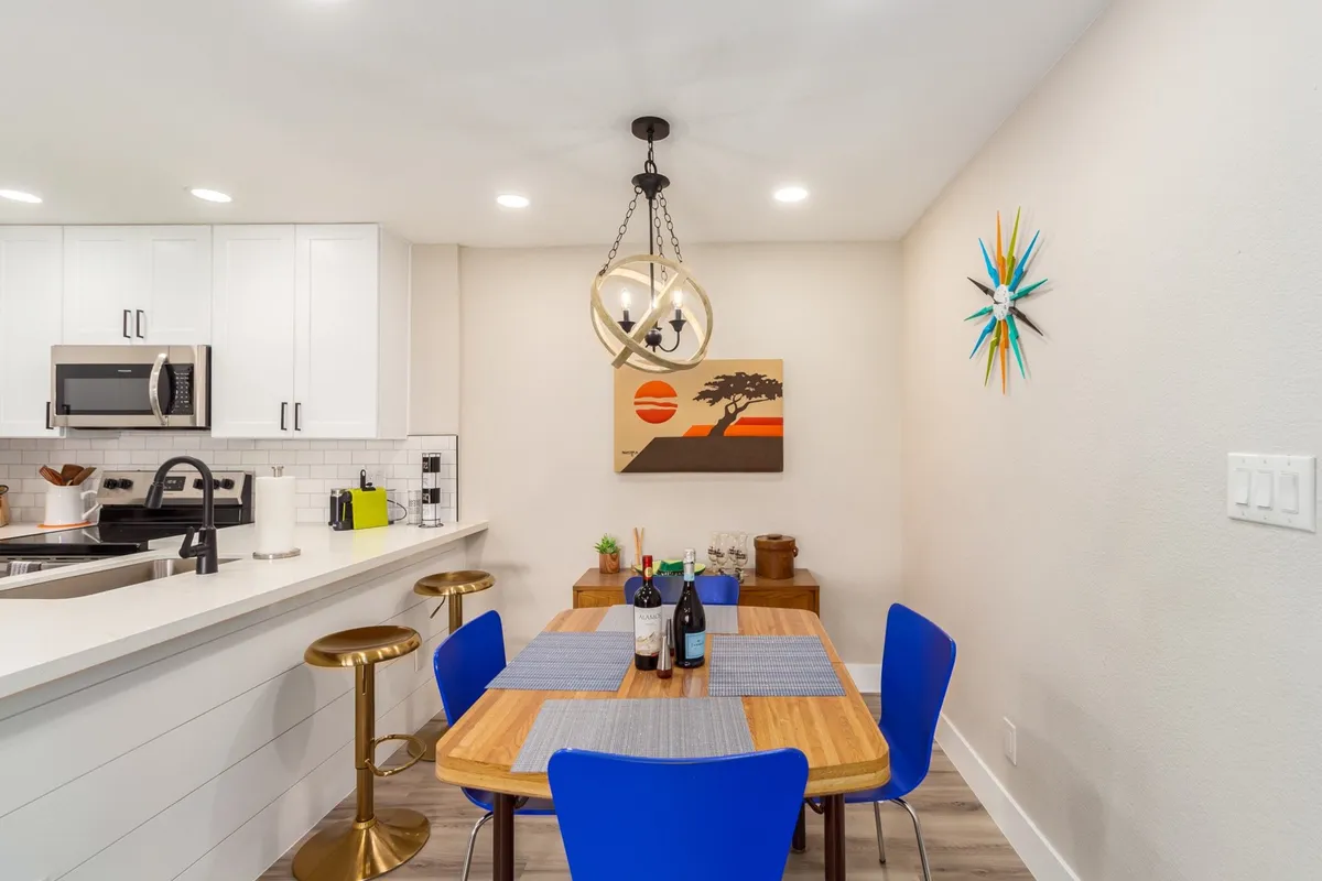 A warmly lit dining area featuring a central dining table, stylish artwork on the wall, and bar stools lining a counter, set up for both meals and gatherings. The scene is inviting, with enough space for both dining and work tasks.