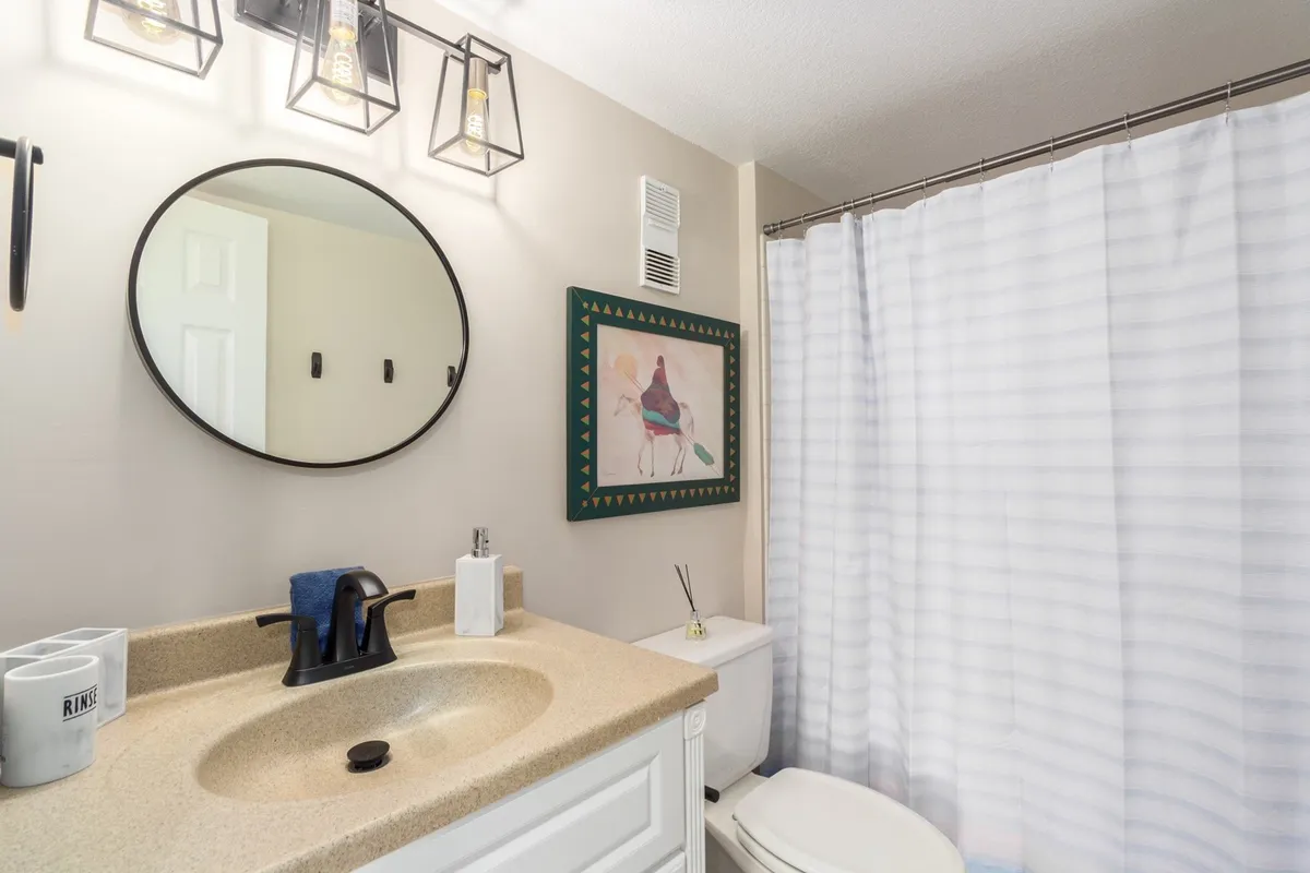 A serene bathroom scene with a round mirror, modern sink area, stylish lighting fixtures, and soothing wall art. The neutral tones and soft lighting evoke a sense of calm and personal retreat.