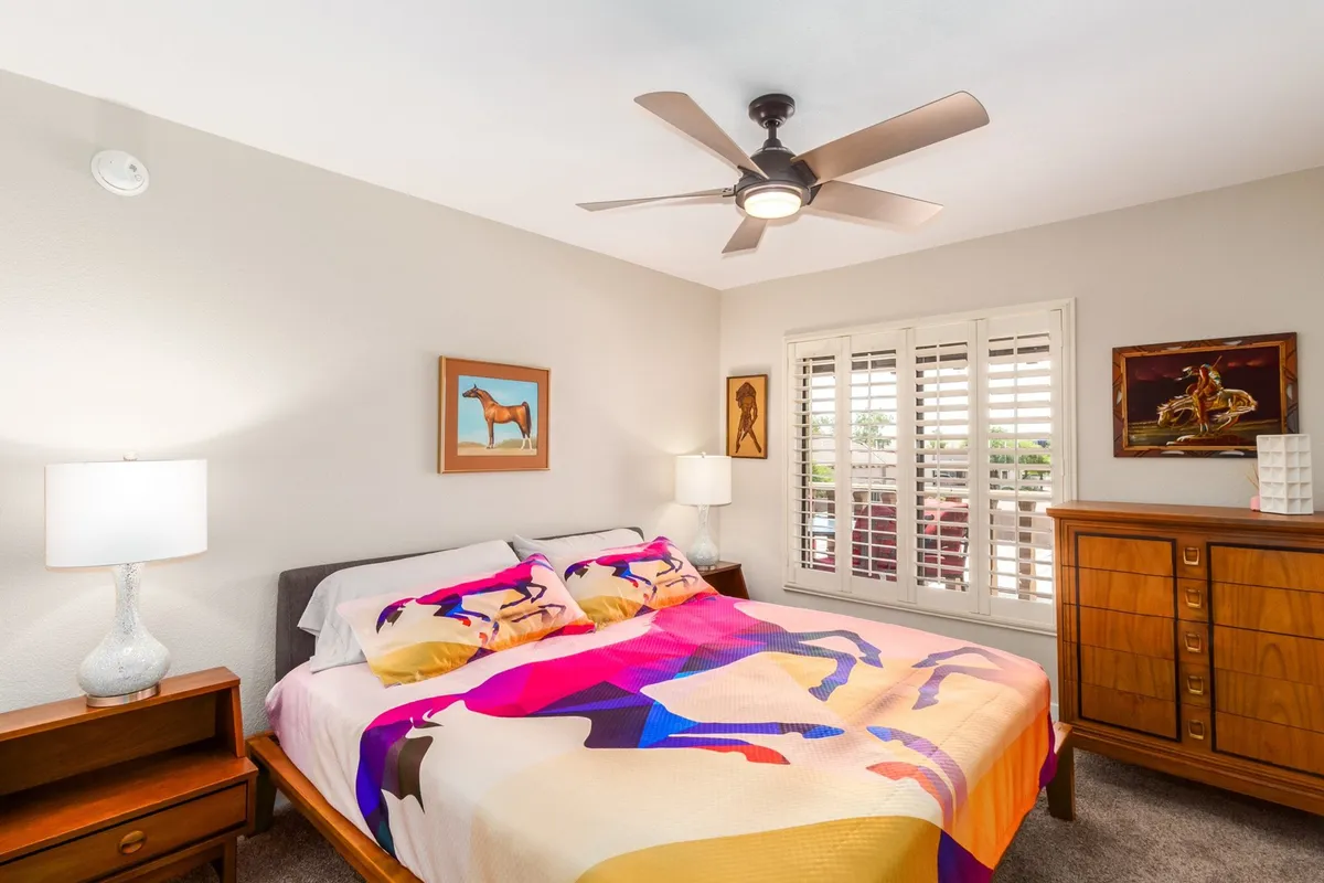 This image features a cozy bedroom with a colorful bedspread, artwork on the walls, and warm wooden furniture. Sunlight softly fills the room, creating a tranquil and inviting atmosphere ideal for relaxation or quiet moments.