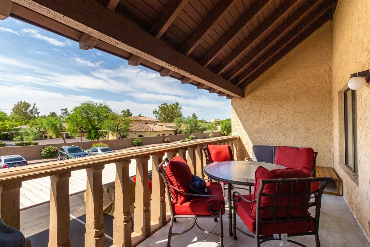 This image features a spacious balcony setup with red cushioned chairs, a round table, classic balcony railing, and a lush backdrop of trees and distant buildings. The space is set for outdoor relaxation or dining, with plenty of seating, and a calm, inviting atmosphere that aligns with tranquil Arizona evenings.
