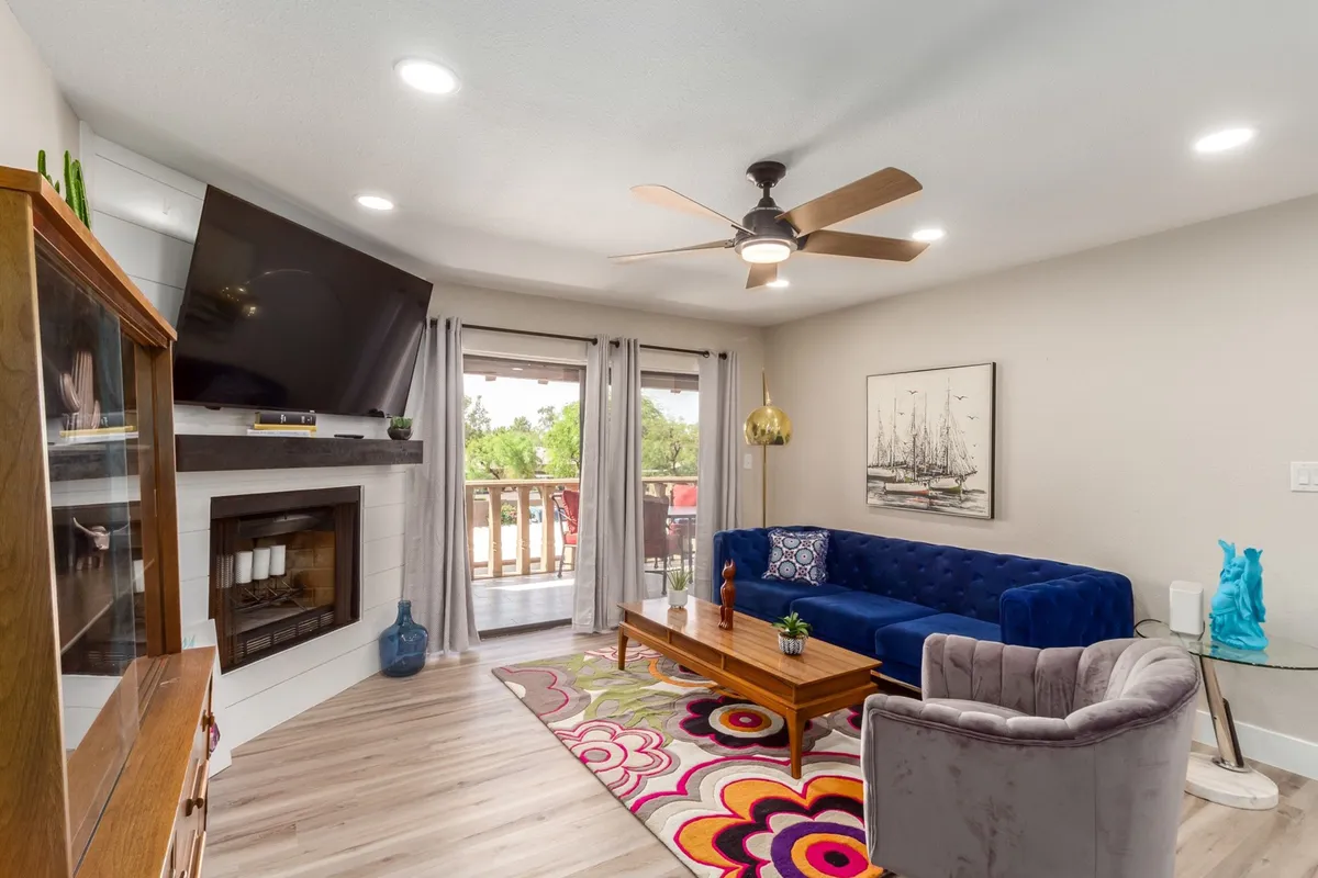 A cheerful, inviting living area featuring a plush blue velvet sofa, a vibrant colorful rug, a wooden coffee table, a glowing fireplace, and a large television. The space is modern and cozy, ideal for relaxing, socializing, or enjoying a movie night.