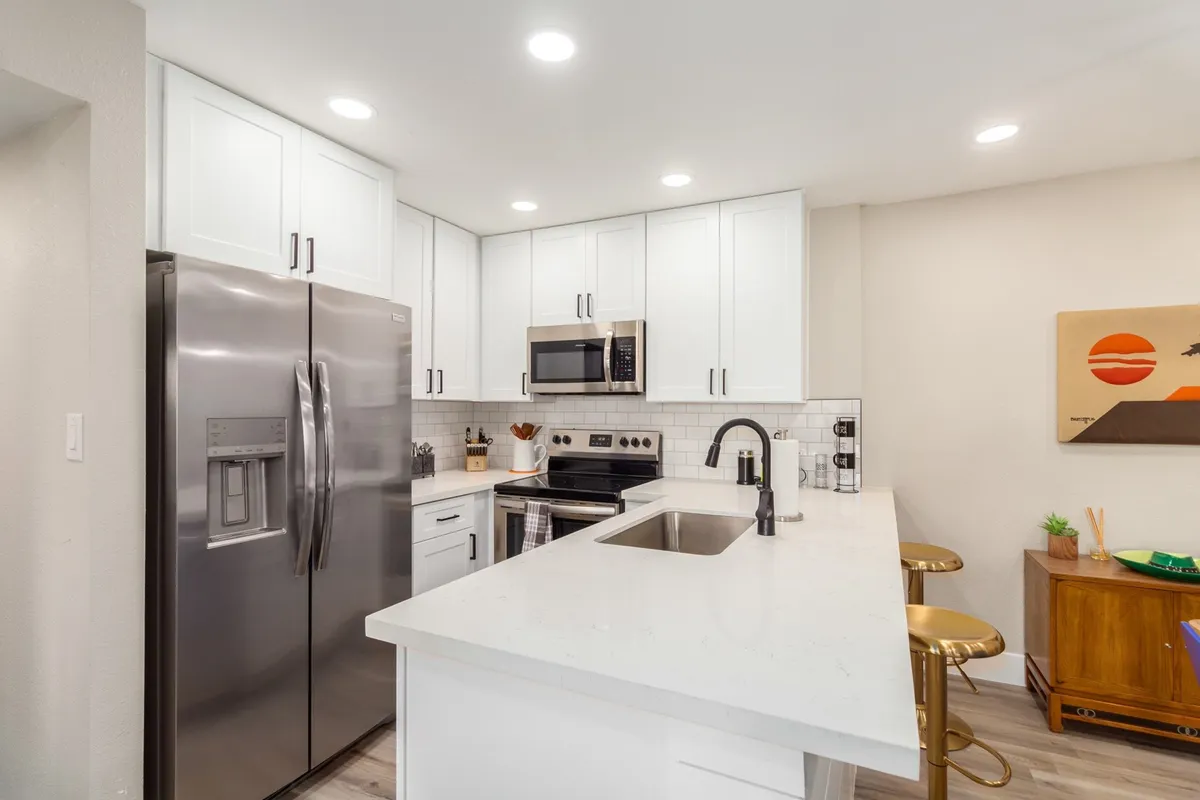 A bright, contemporary kitchen highlighted by a sleek stainless steel refrigerator, expansive quartz countertops, a central island with seating, eye-catching modern cabinetry, and decorative artwork. The space exudes a welcoming, high-end vibe for both cooking and casual conversation.