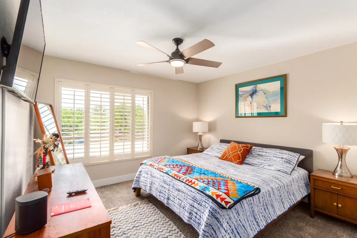 A cozy bedroom with a king-sized bed dressed in a vibrant blanket, framed artwork above the headboard, shuttered windows that let in natural light, and a nightstand with a lamp. The space feels intimate and inviting, perfect for relaxation.