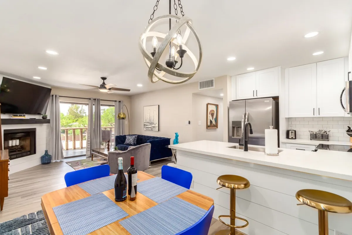 This image captures an open-concept space that merges the dining table with blue chairs, a kitchen island, a spacious living room seating area, a cozy fireplace, and a large TV. The area is filled with natural light and lively accents, emphasizing the seamless layout and versatility for both entertaining and everyday living.