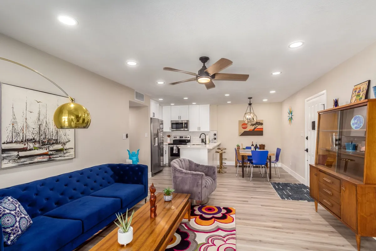 A cheerful, open-plan living and dining space with a blue tufted sofa, a wooden coffee table, a colorful rug, and a modern dining area. The scene showcases an area ideal for relaxing after a busy day, with plenty of natural light and inviting decor.