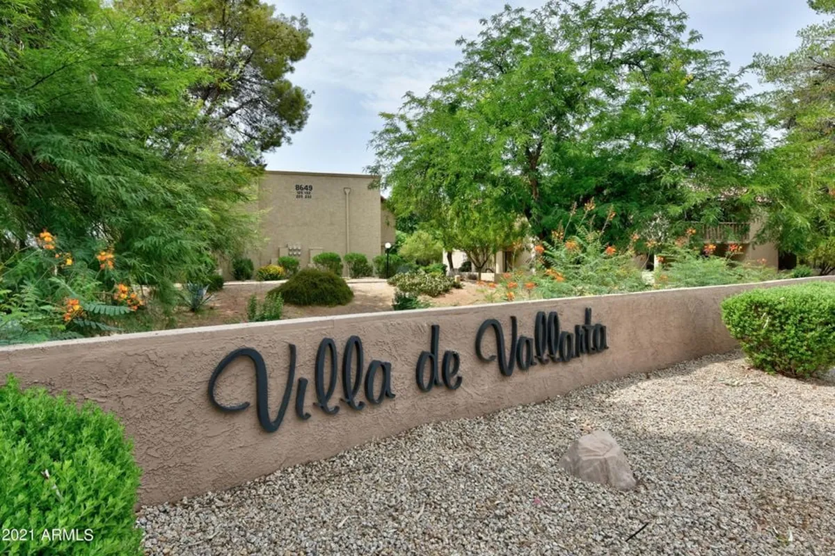 The entrance to Villa de Vallarta is framed by lush greenery, decorative stones, and a welcoming sign. The scene feels peaceful and inviting, with pathways ideal for leisurely walks and an abundance of seasonal flowers enhancing the vibrant atmosphere.